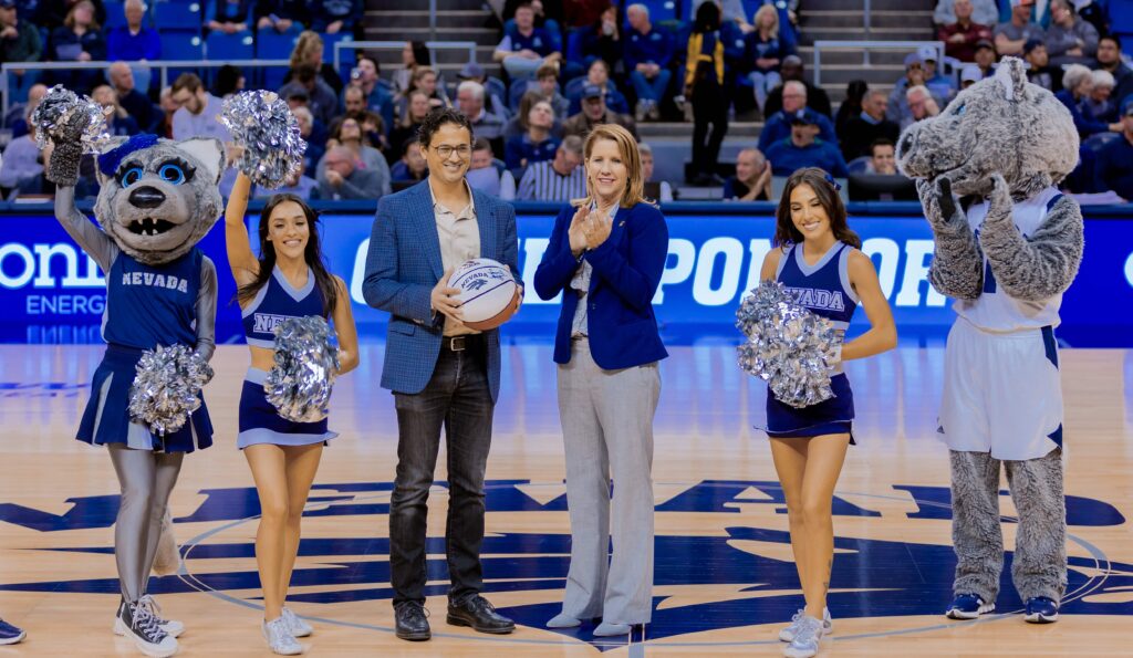 CEO of Dragonfly Energy, Denis Phares is smiling on the basketball court of the Lawlor Events Center. He holds a University of Nevada branded basketball and has cheerleaders and mascots on both sides of him.